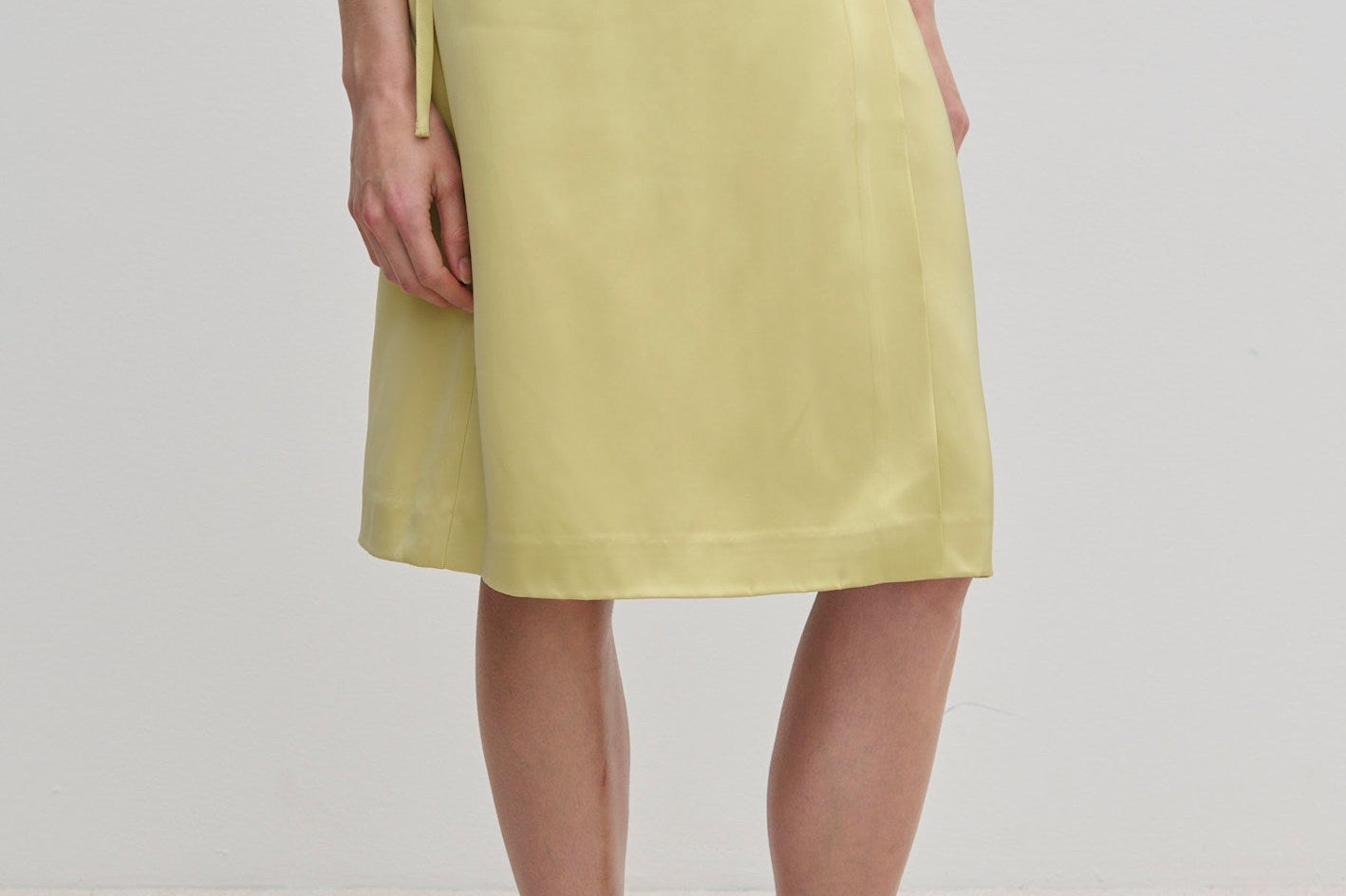 A person wearing a pale yellow knee-length Emmy Wrap Skirt by Herskind ApS, featuring an A-line silhouette tied at the waist with a simple side bow and black flat shoes.