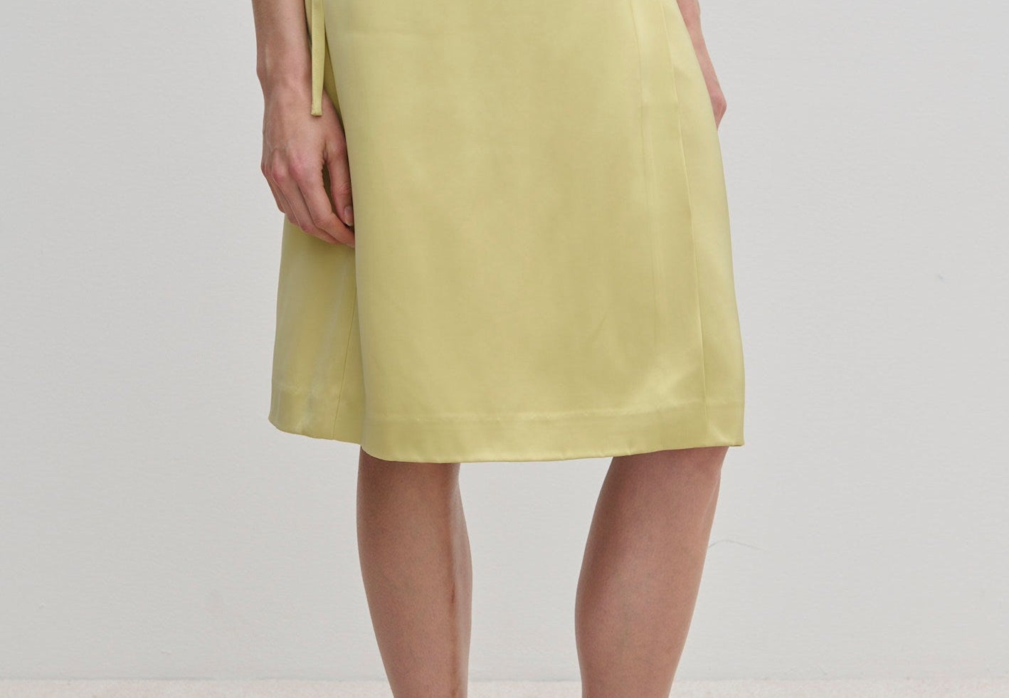 A person wearing a pale yellow knee-length Emmy Wrap Skirt by Herskind ApS, featuring an A-line silhouette tied at the waist with a simple side bow and black flat shoes.