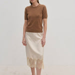 A woman wearing a brown Thora Knit Blouse in wool and cashmere, a cream-colored midi skirt with a lace hem, and black heeled thong sandals.