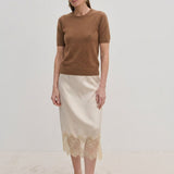 A woman wearing a brown Thora Knit Blouse in wool and cashmere, a cream-colored midi skirt with a lace hem, and black heeled thong sandals.