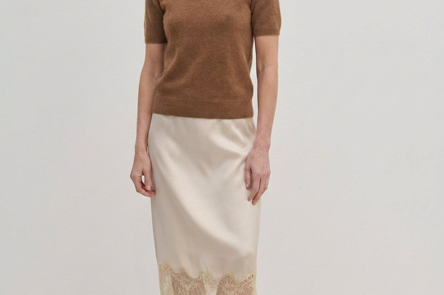 A woman wearing a brown Thora Knit Blouse in wool and cashmere, a cream-colored midi skirt with a lace hem, and black heeled thong sandals.