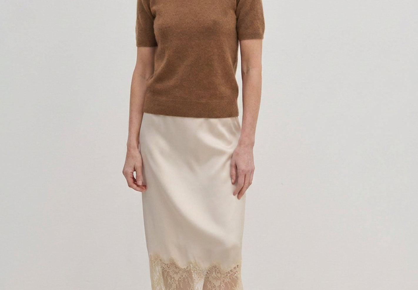 A woman wearing a brown Thora Knit Blouse in wool and cashmere, a cream-colored midi skirt with a lace hem, and black heeled thong sandals.