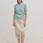 A woman wearing a pale green Thora Knit Blouse by Herskind ApS and a beige satin midi skirt with a lace hem, paired with black heeled sandals.