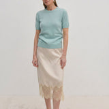 A woman wearing a pale green Thora Knit Blouse by Herskind ApS and a beige satin midi skirt with a lace hem, paired with black heeled sandals.