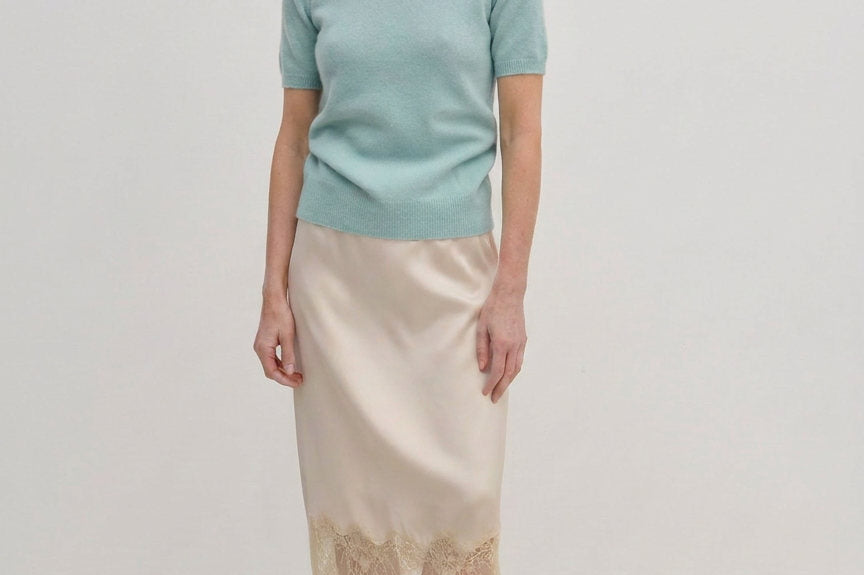 A woman wearing a pale green Thora Knit Blouse by Herskind ApS and a beige satin midi skirt with a lace hem, paired with black heeled sandals.