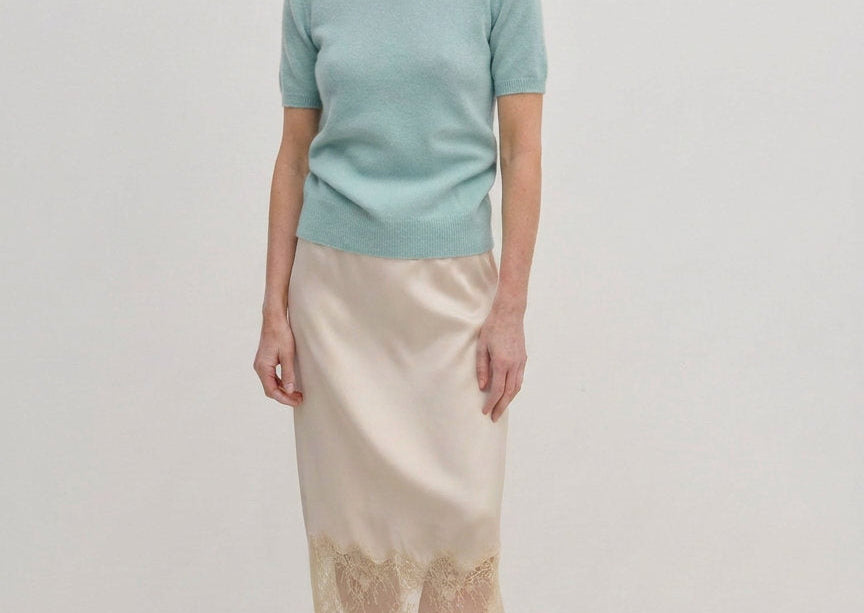 A woman wearing a pale green Thora Knit Blouse by Herskind ApS and a beige satin midi skirt with a lace hem, paired with black heeled sandals.