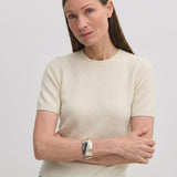 A woman wears a cream-colored, short-sleeved crew-neck Thora Knit Blouse by Herskind ApS, paired with grey trousers and a black belt.