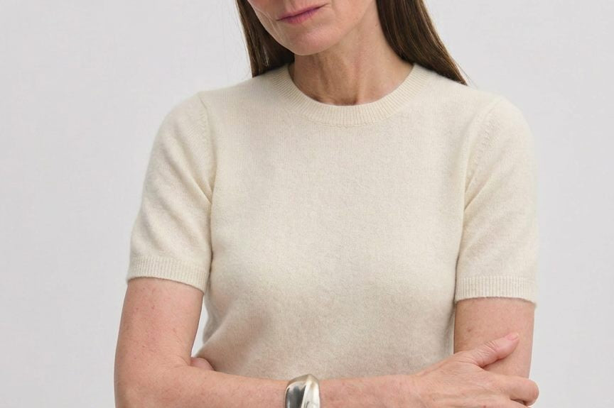 A woman wears a cream-colored, short-sleeved crew-neck Thora Knit Blouse by Herskind ApS, paired with grey trousers and a black belt.