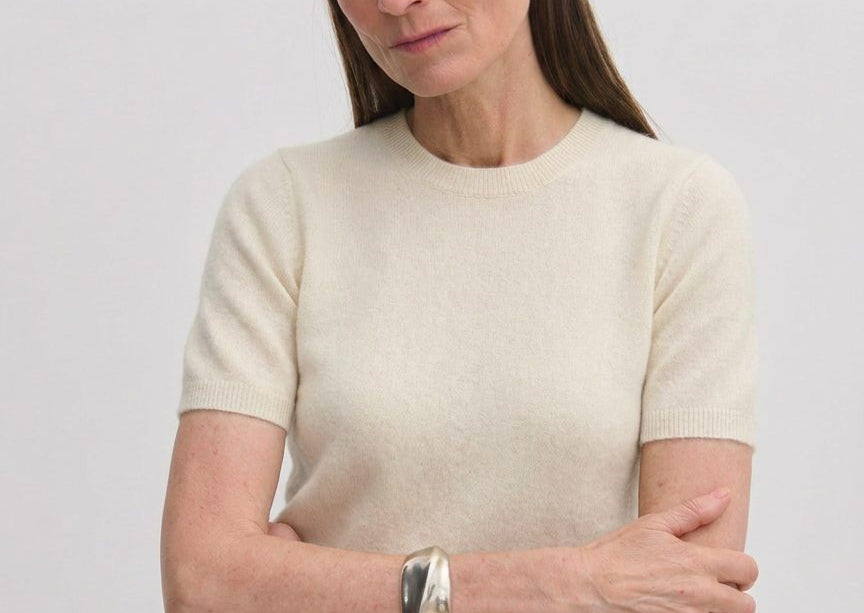 A woman wears a cream-colored, short-sleeved crew-neck Thora Knit Blouse by Herskind ApS, paired with grey trousers and a black belt.