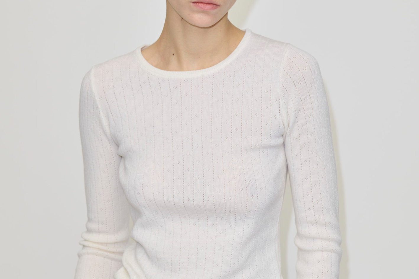 A person wearing the Herskind ApS Camb Knit Blouse, a long-sleeved, white merino wool ribbed knit top with pointelle detailing.