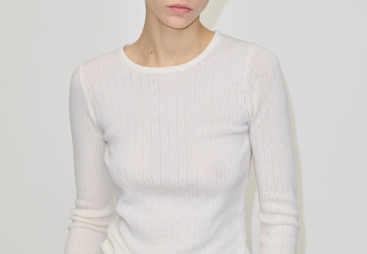 A person wearing the Herskind ApS Camb Knit Blouse, a long-sleeved, white merino wool ribbed knit top with pointelle detailing.