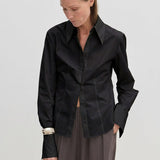 A slim-fit, long-sleeved black Vivienne Shirt by Herskind ApS with a pointed collar and wide cuffs, worn over grey trousers.