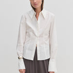 A person wearing a Vivienne Shirt by Herskind ApS, a slim fit, white, long-sleeved button-down with a pointed collar and wide cuffs, paired with dark grey trousers.