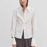 A person wearing a Vivienne Shirt by Herskind ApS, a slim fit, white, long-sleeved button-down with a pointed collar and wide cuffs, paired with dark grey trousers.