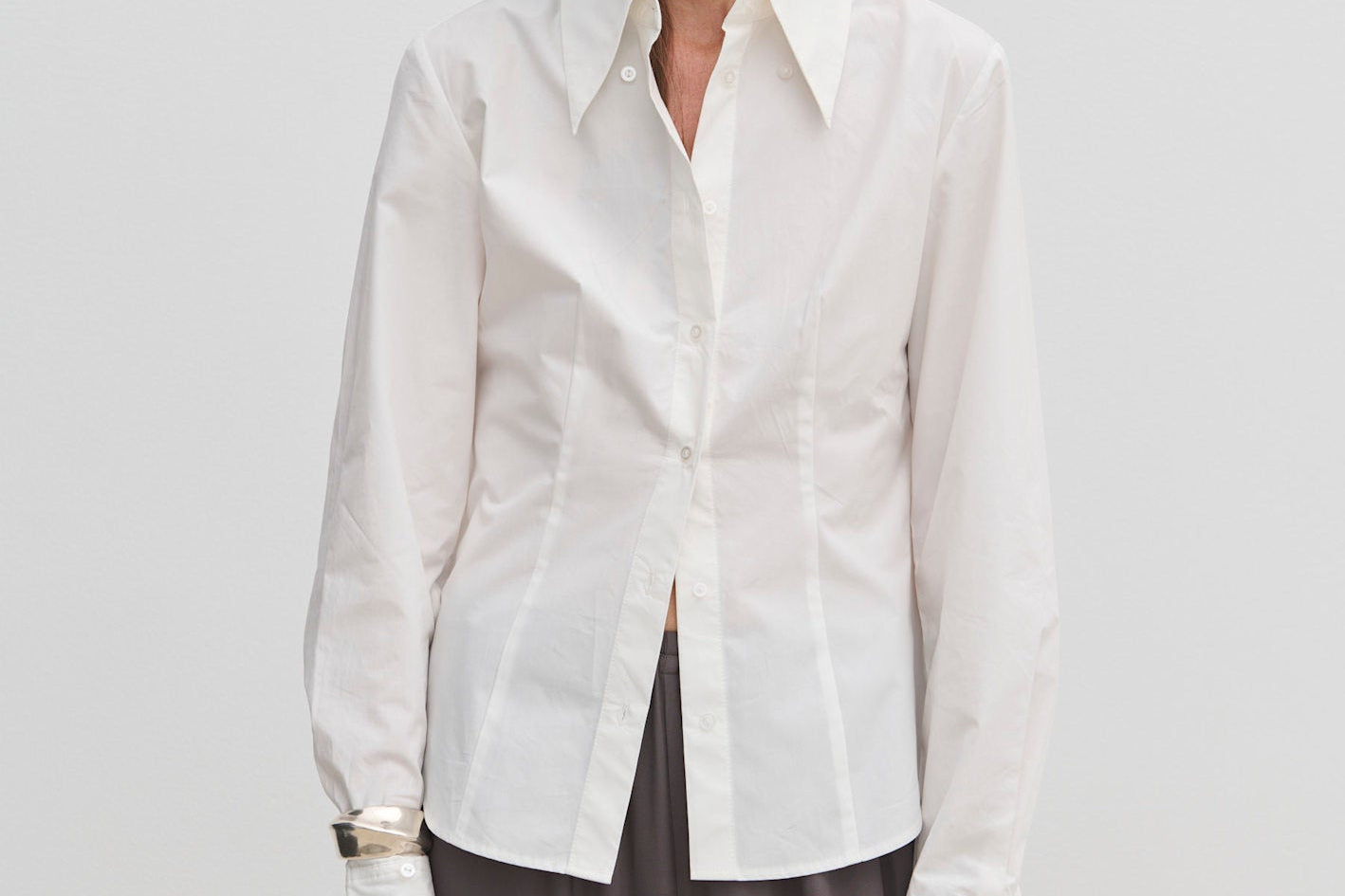A person wearing a Vivienne Shirt by Herskind ApS, a slim fit, white, long-sleeved button-down with a pointed collar and wide cuffs, paired with dark grey trousers.