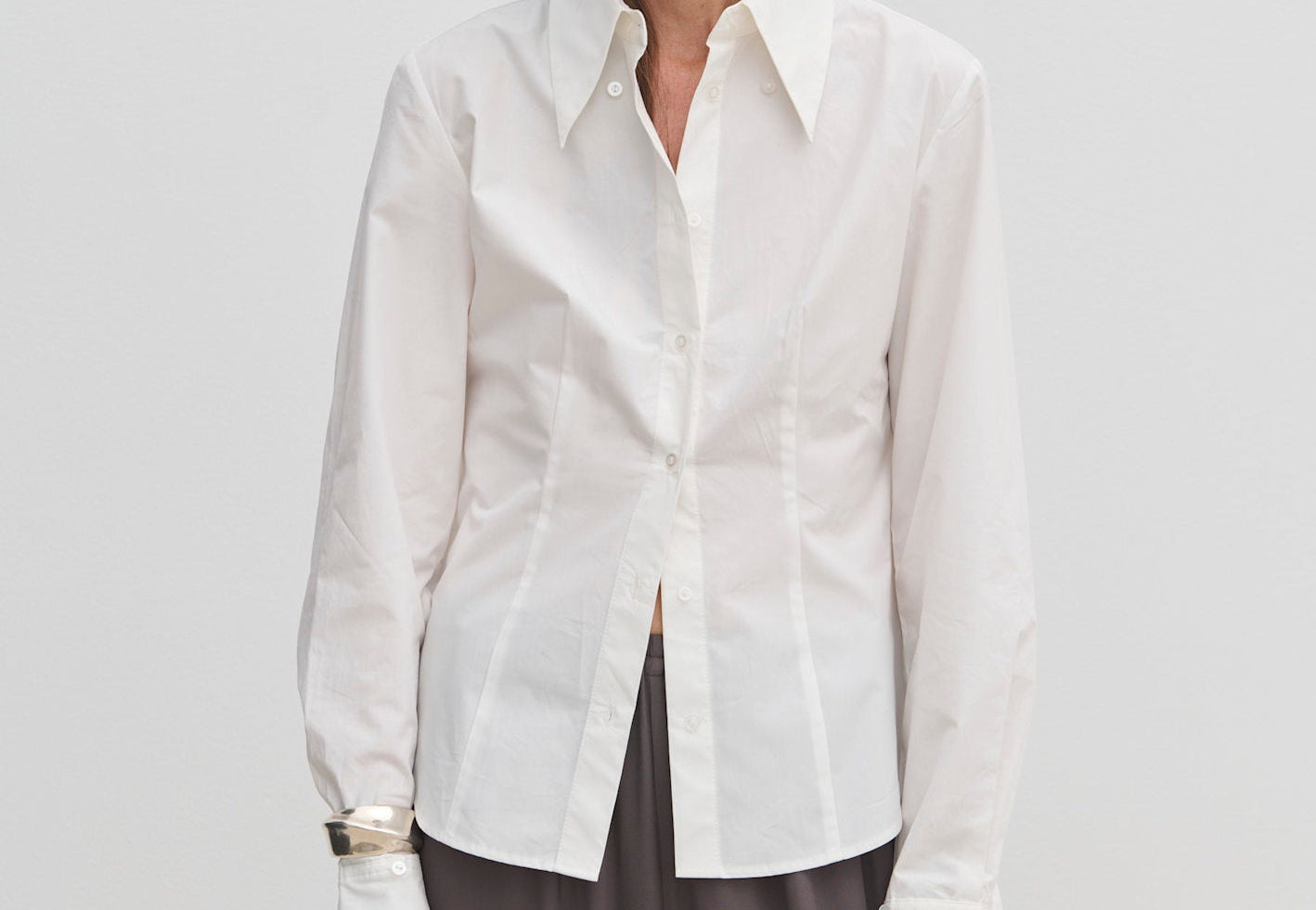 A person wearing a Vivienne Shirt by Herskind ApS, a slim fit, white, long-sleeved button-down with a pointed collar and wide cuffs, paired with dark grey trousers.