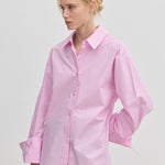 A woman wears an oversized light pink cotton Inga Shirt by Herskind ApS with wide, folded cuffs, paired with grey trousers.