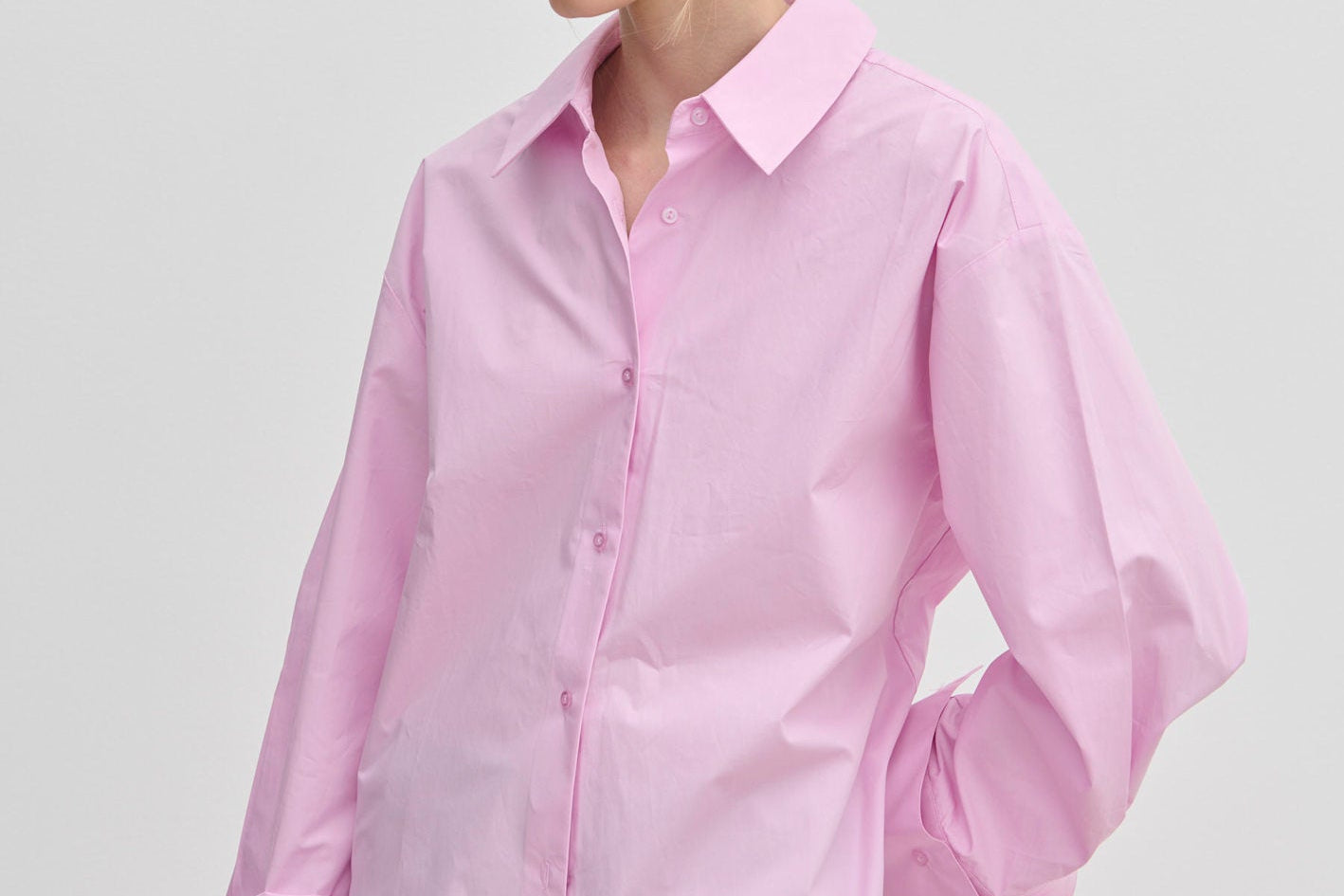 A woman wears an oversized light pink cotton Inga Shirt by Herskind ApS with wide, folded cuffs, paired with grey trousers.
