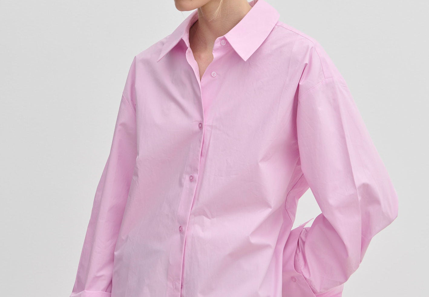 A woman wears an oversized light pink cotton Inga Shirt by Herskind ApS with wide, folded cuffs, paired with grey trousers.