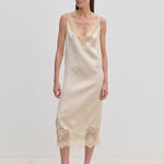 A woman wears a champagne-colored Florence Dress by Herskind ApS, a sleeveless midi slip dress with lace trim at the neckline and hem, paired with beige pointed-toe heels.