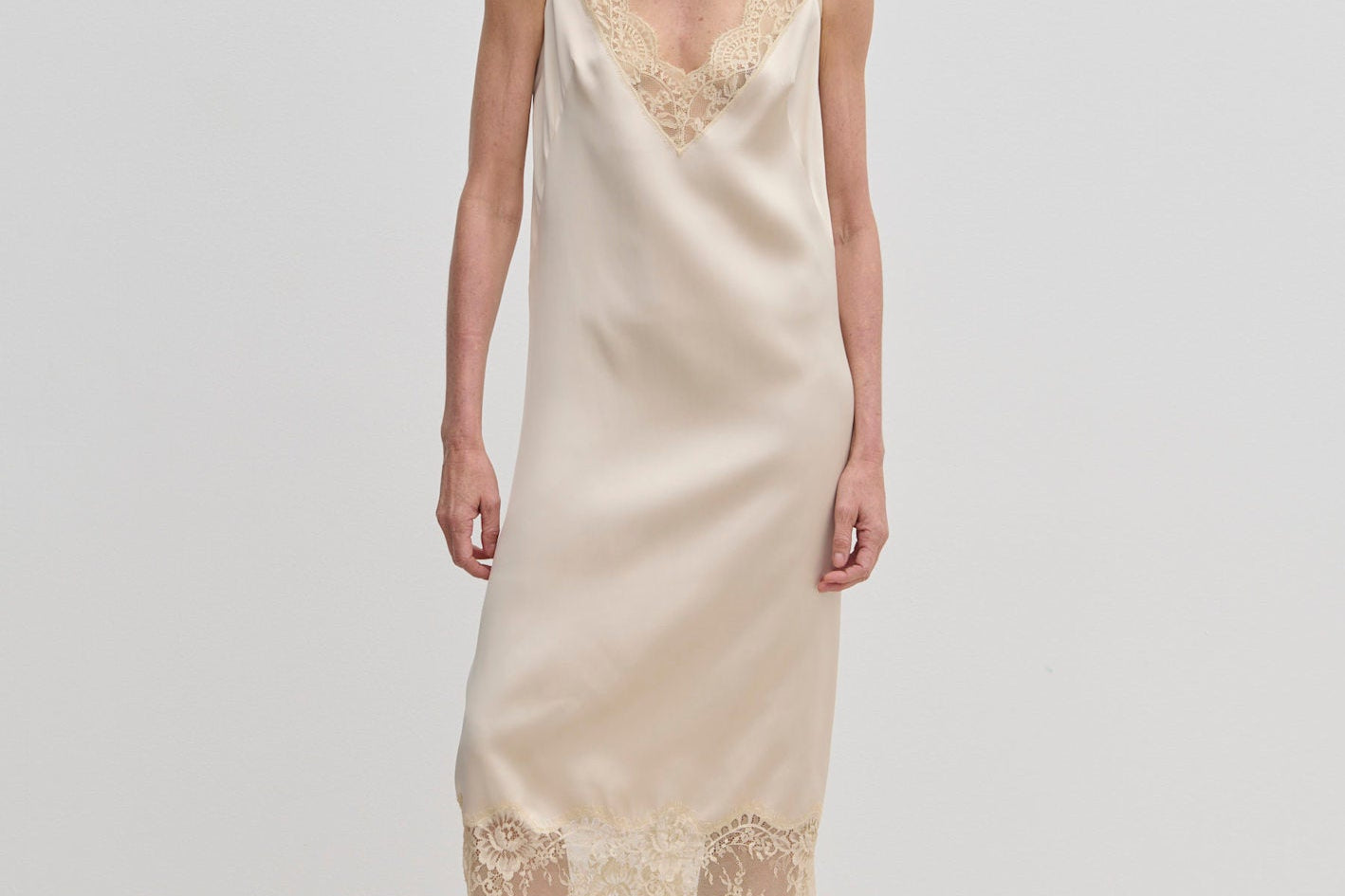 A woman wears a champagne-colored Florence Dress by Herskind ApS, a sleeveless midi slip dress with lace trim at the neckline and hem, paired with beige pointed-toe heels.