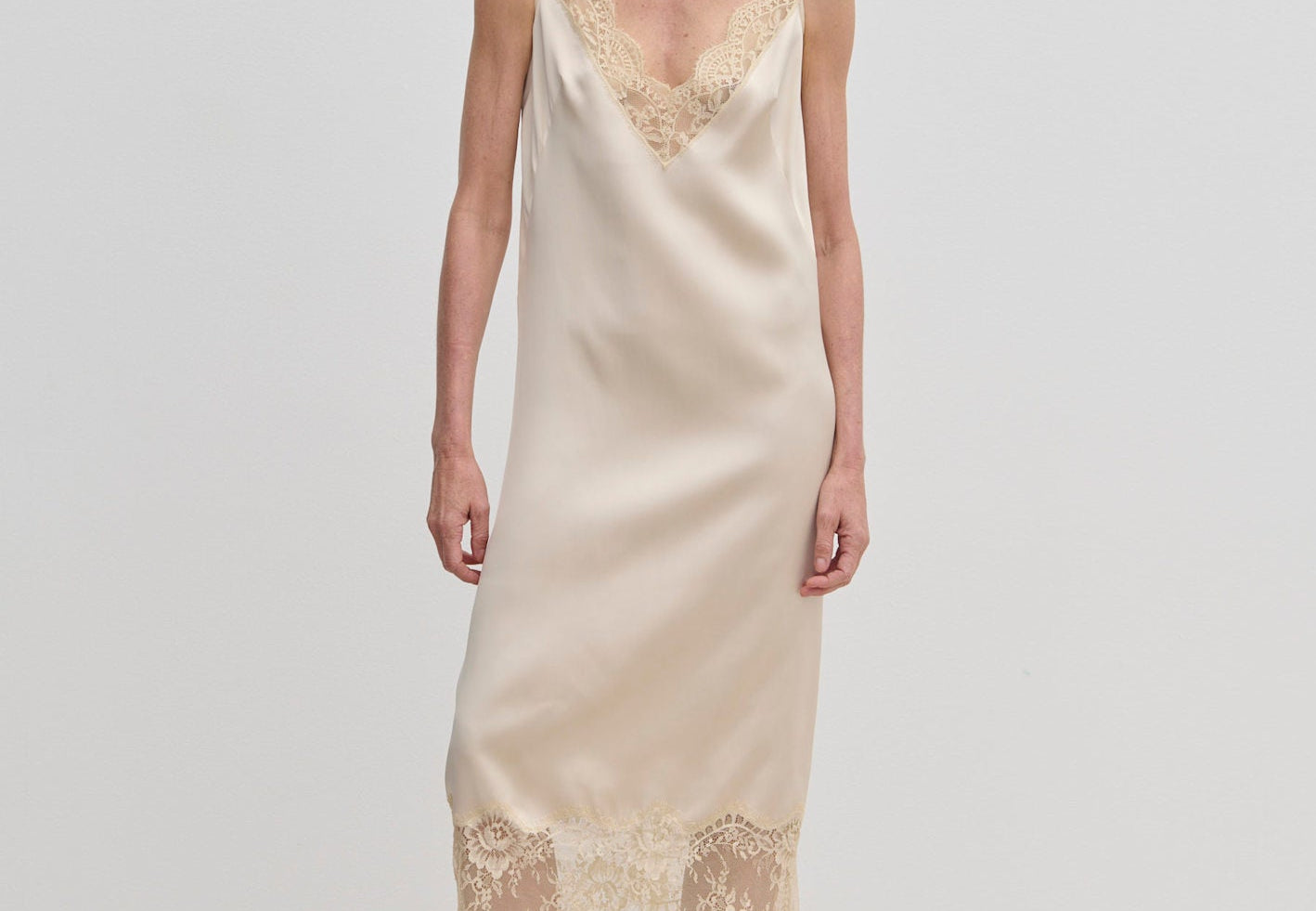A woman wears a champagne-colored Florence Dress by Herskind ApS, a sleeveless midi slip dress with lace trim at the neckline and hem, paired with beige pointed-toe heels.