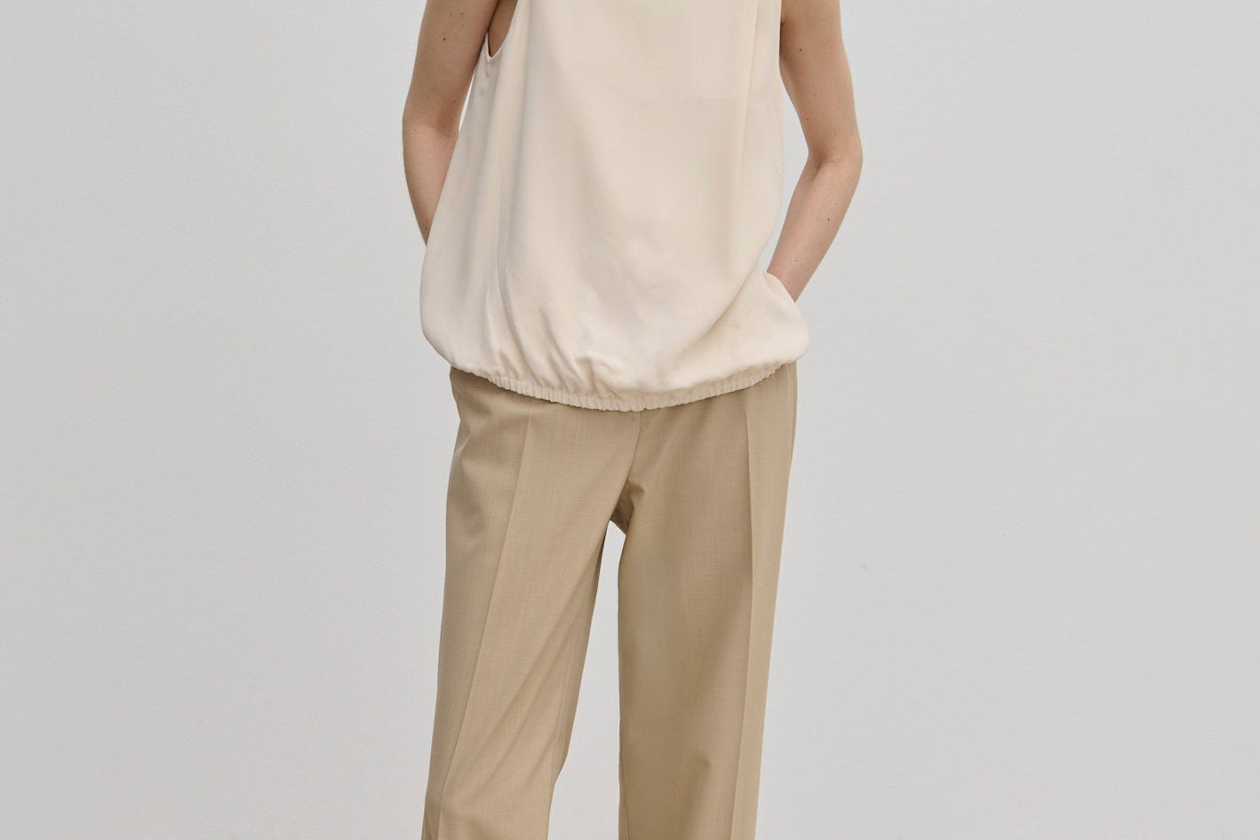 A model wearing a cream-colored Loa Top by Herskind ApS with a sleeveless, high funnel neck and a cinched hem, paired with tan trousers and cream-colored shoes.