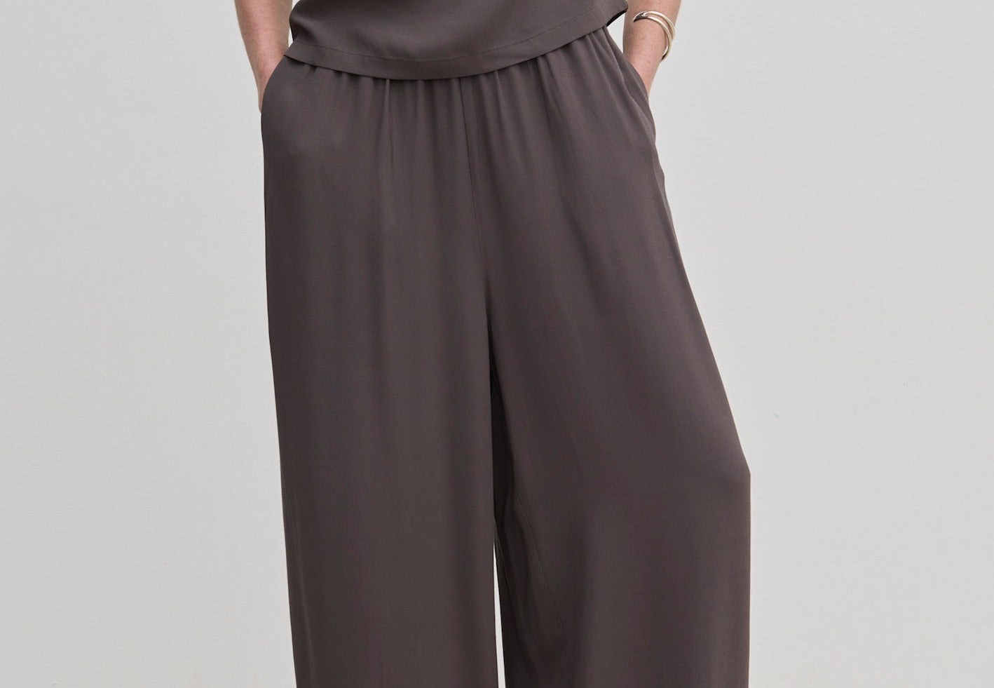 A person wears dark gray Nya Pants by Herskind ApS, featuring balloon legs, an elasticated waist, and a draped top.