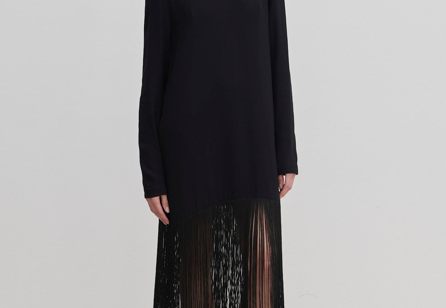 The Aris Dress by Herskind ApS is a long-sleeved black shift dress with a crew neckline and an ankle-length fringe hem, styled with black mules.