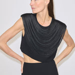 A woman wears a black, cropped Dita Top by Herskind ApS, featuring a sleeveless design with draped fringe layers across the chest and a midriff cut-out.