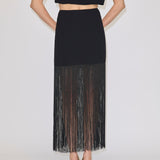 The Herskind ApS Olivia Skirt is a black high-waisted midi skirt with long, sheer fringe at the hem, worn with a matching sleeveless top and cream heels.