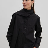 A person wears an oversized black cotton Natalia Shirt by Herskind ApS with an attached scarf-like neck detail and tan trousers.