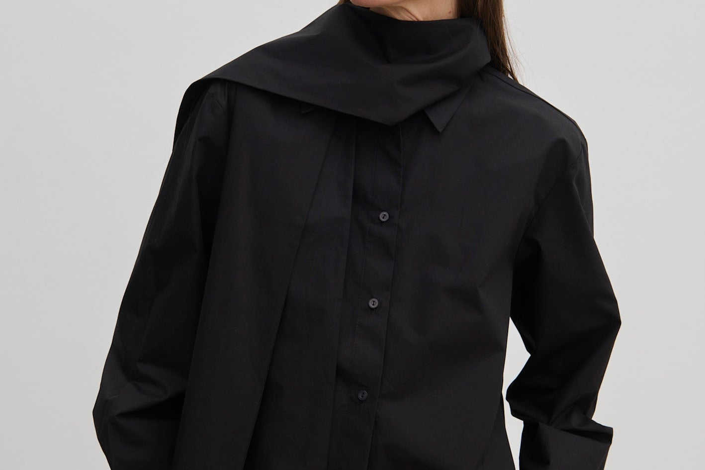 A person wears an oversized black cotton Natalia Shirt by Herskind ApS with an attached scarf-like neck detail and tan trousers.