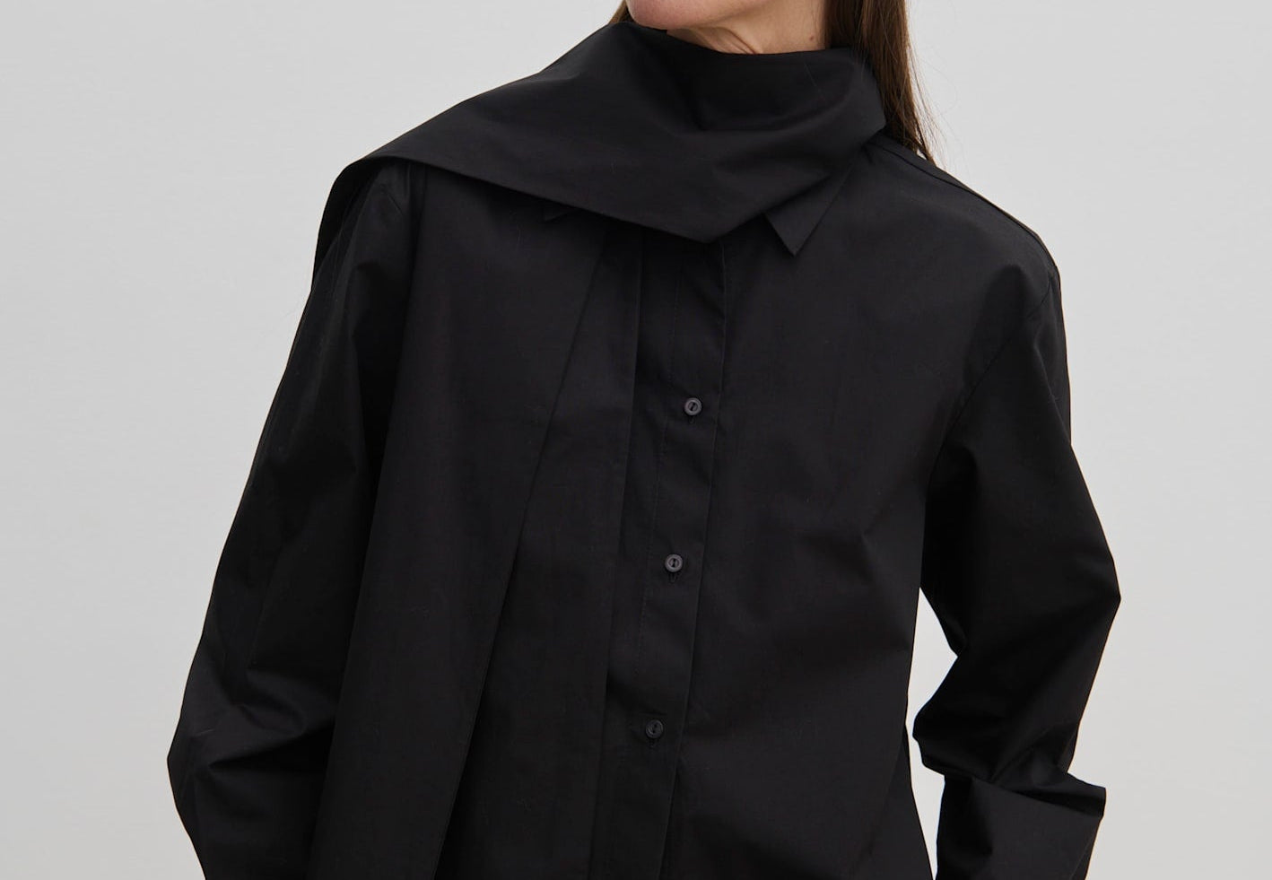 A person wears an oversized black cotton Natalia Shirt by Herskind ApS with an attached scarf-like neck detail and tan trousers.