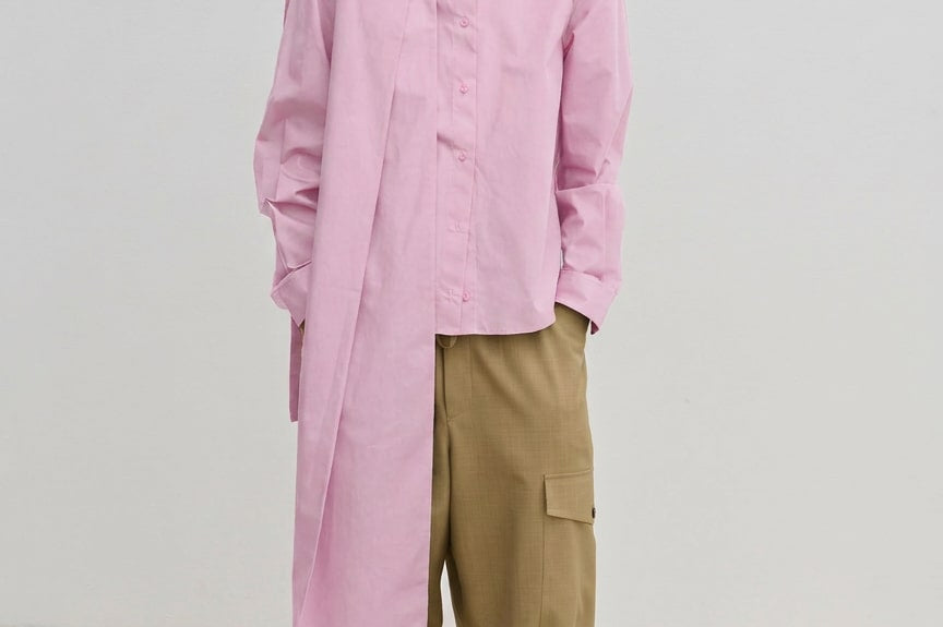 A person wears the Herskind ApS Natalia Shirt, an oversized, long-sleeved light pink button-down with a detachable scarf panel at the shoulder, paired with wide-leg tan cargo pants.