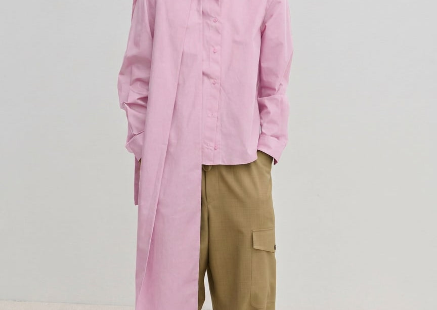A person wears the Herskind ApS Natalia Shirt, an oversized, long-sleeved light pink button-down with a detachable scarf panel at the shoulder, paired with wide-leg tan cargo pants.